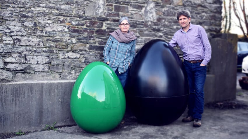The image shows Fiona Kelleher and Kieran Coffey standing outdoors against a stone wall, each positioned beside large, egg-shaped MyGug. On the left, Fiona, wearing a plaid shirt and scarf, stands next to a shiny green MyGug unit. On the right, Kieran in a purple checkered shirt with his hands in his pockets leans against a black MyGug unit. Both are smiling.