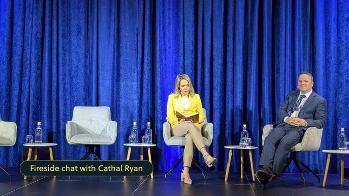 A woman in a yellow blazer and beige trousers sits on stage reading from a notebook, while a man in a dark suit and blue tie sits beside her, smiling. They are part of a panel setup with empty chairs and water bottles, in front of a backdrop of deep blue curtains. A caption at the bottom reads, “Fireside chat with Cathal Ryan.”