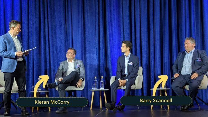 A panel discussion on stage with four men in business attire seated in front of blue curtains. The man on the far left is standing and holding a notebook, while the three seated participants are engaged in conversation. Kieran McCorry is second from the left, smiling with legs crossed, and Barry Scannell is on the far right. Arrows and name labels identify them at the bottom of the image.
