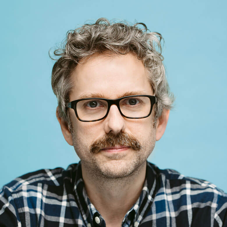 portrait of a man with curly grey hair, glasses, and a moustache wearing a blue checked shirt, against a light blue background.