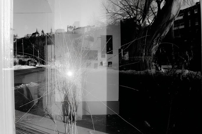 Black-and-white photograph showing a street scene reflected through cracked and scratched glass, with a bright glare near the center, bare trees, snow along the roadside, and overlapping reflections of buildings that partially obscure the view.
