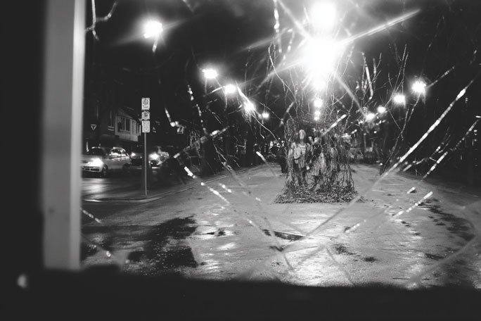 Black-and-white nighttime street scene viewed through cracked and shattered glass, with bright streetlights reflecting off a wet road, cars along the sides, and a small group of people standing together in the center distance, partially obscured by branching cracks across the frame.