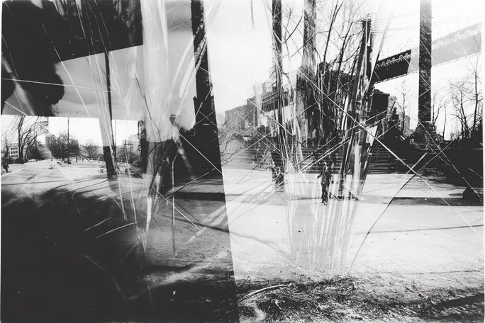 Black-and-white composite photograph of an outdoor urban space, with overlapping panels of a snowy open area, bare trees, and distant buildings, all seen through heavily cracked and scratched glass that creates vertical splits and jagged lines across the scene, partially obscuring two small figures walking in the center.