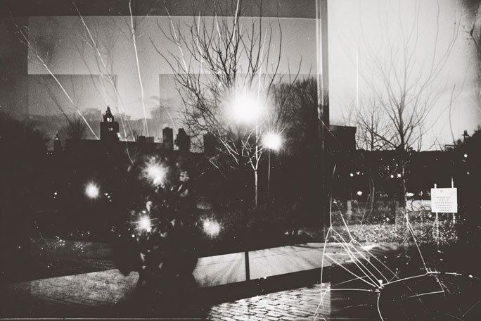 Black-and-white photograph showing an urban park or plaza at dusk, layered with reflections and scratches from glass, with leafless trees silhouetted against the sky, bright circular light flares scattered across the scene, a cobblestone path in the foreground, and distant buildings and signage partially obscured by overlapping panels and crack lines.
