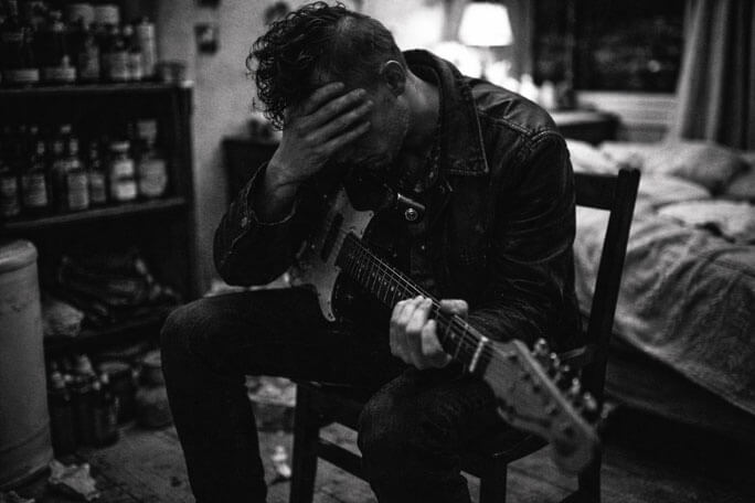 Black-and-white scene of a person seated on a chair indoors, holding an electric guitar across their lap while covering their face with one hand. Their posture is slumped, suggesting distress or exhaustion. A bed with rumpled bedding is behind them, and shelves filled with jars or bottles line the wall, creating an intimate, cluttered room atmosphere. AI generated.