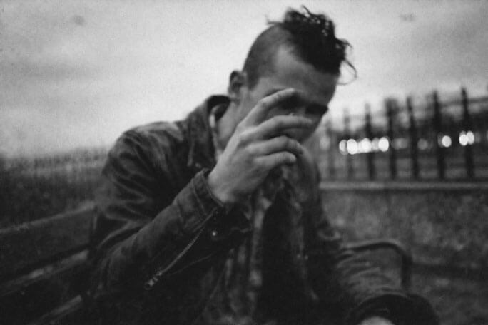 Black-and-white photograph of a person seated on a bench outdoors, with short sides and a tousled strip of hair on top, leaning forward and covering part of their face with one hand, with a metal fence and blurred city lights in the background.