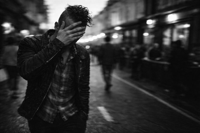 Black-and-white photograph of a person walking alone down a busy city street at night, wearing a jacket and plaid shirt, with one hand covering their face and head tilted downward, while blurred pedestrians and glowing storefront lights recede into the background along a wet roadway.